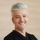 A smiling person with short, styled light hair and a black shirt stands against a neutral background. They exude a friendly and approachable demeanor.