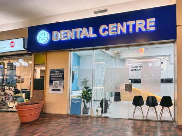 A modern dental center with a bright blue sign reading "DENTAL CENTRE." The glass storefront features a welcoming interior and three black chairs outside. The entrance is located in a shopping area, providing easy access for patients.