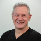 A smiling professional in dark scrubs stands against a light background, conveying a welcoming and friendly demeanor.