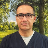 A person wearing navy scrubs and glasses stands against a scenic background of greenery and trees, conveying a professional yet approachable demeanor.