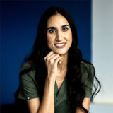 A smiling woman with long black hair rests her chin on her hand, dressed in a green medical uniform, set against a blue background. The image conveys a friendly and approachable demeanor.