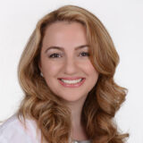 A smiling woman with long, wavy blonde hair is wearing a white coat and looking directly at the camera. She has a warm, friendly expression, suggesting professionalism and approachability.