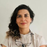 A woman with dark curly hair, wearing a light-colored blouse, smiles at the camera. She has glasses hanging around her neck and stands against a plain background, exuding a professional yet approachable demeanor.