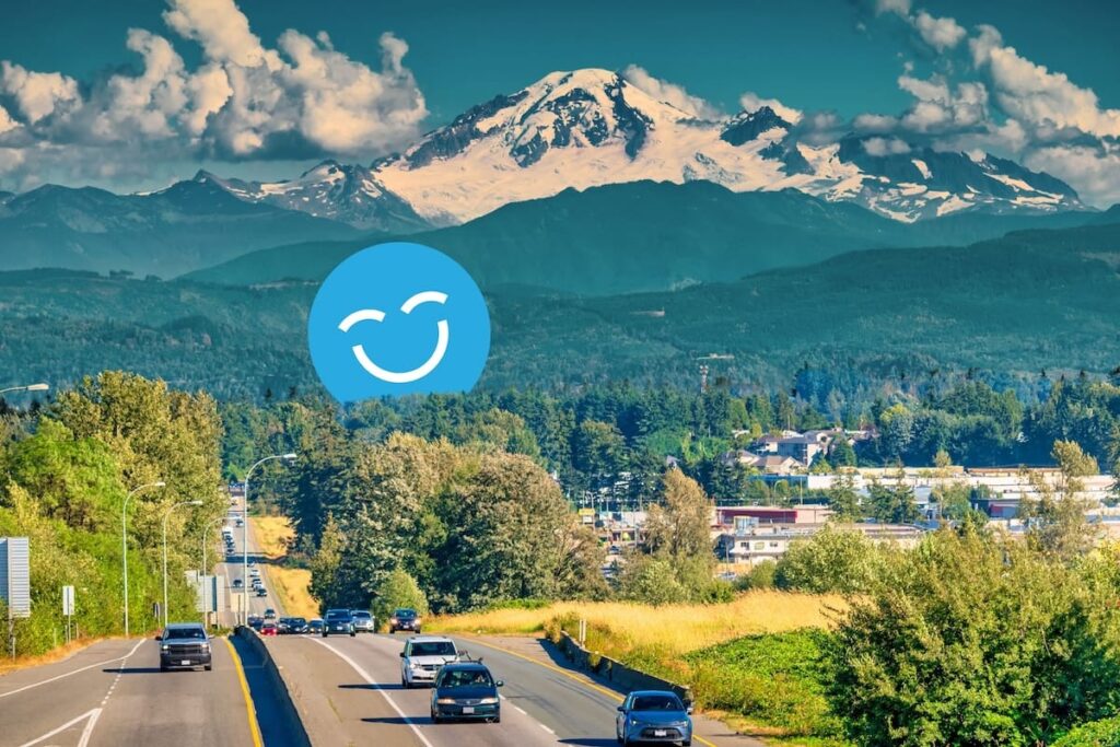 Cars drive on a road with lush greenery on both sides, leading toward a snow-capped mountain under a sky with fluffy clouds.