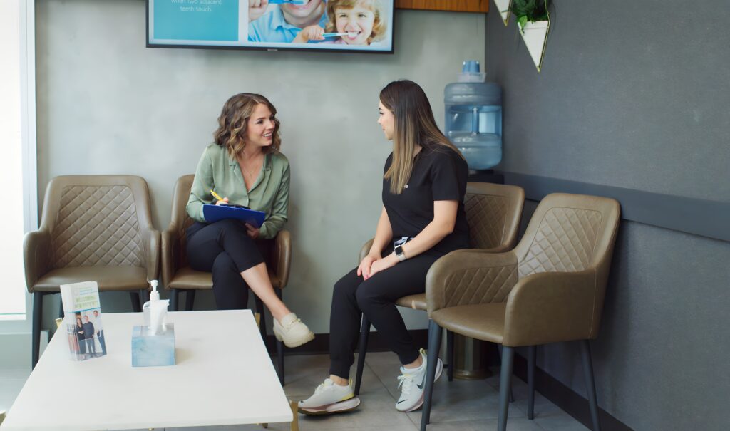 Two women are sitting in a waiting area, engaged in conversation. One woman holds a clipboard, while the other listens attentively. The space features modern furniture and a water cooler in the background.