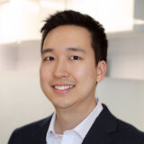 A smiling person in a professional attire poses in an office setting, conveying a friendly and approachable demeanor.