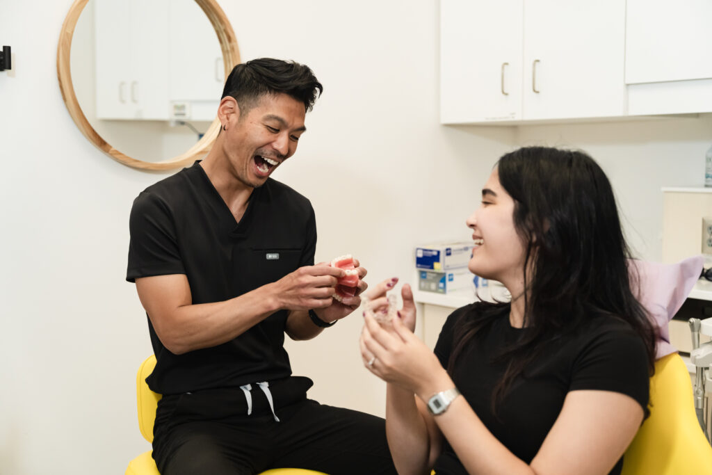 A smiling dental professional demonstrates a product to a laughing patient in a bright, modern office setting. The mood is friendly and engaging, highlighting a positive interaction between them.