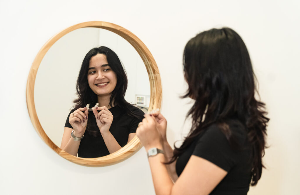 A woman with long hair smiles at her reflection in a round, wooden-framed mirror, holding small objects in her hands. She is wearing a black shirt and appears to be in a bright, minimalistic setting.