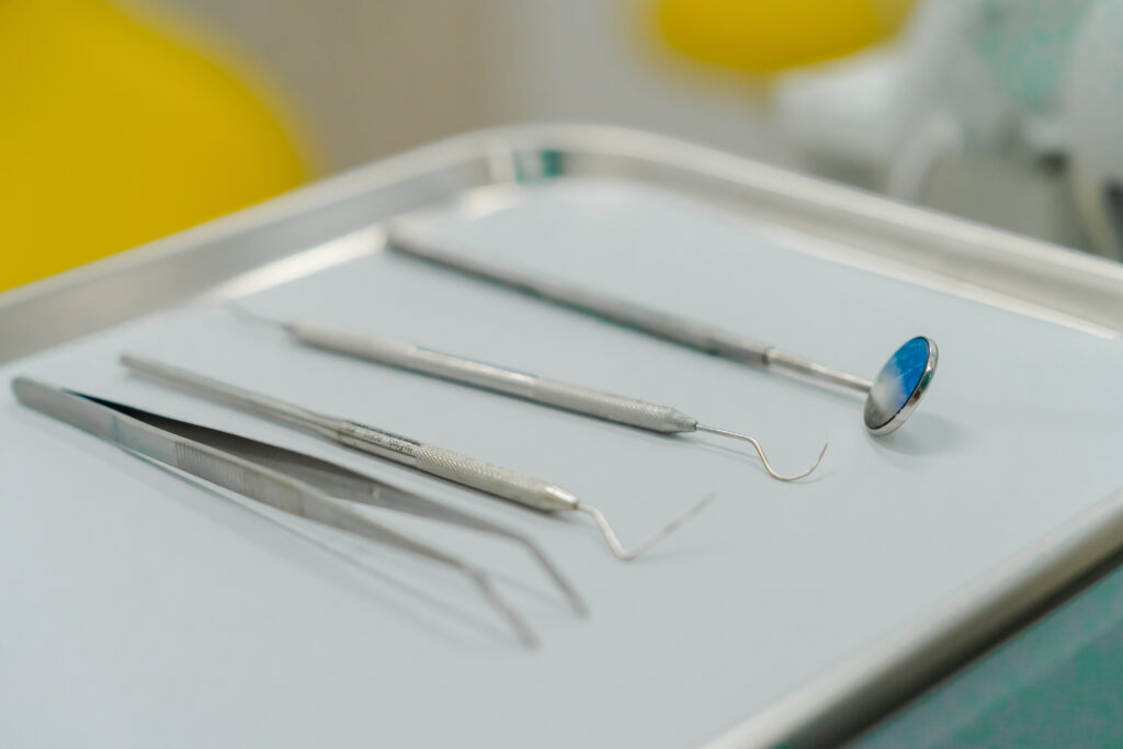 A set of dental tools, including tweezers, a dental mirror, and explorer instruments, arranged on a tray in a dental office setting.