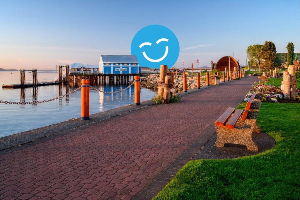 A scenic waterfront path lined with benches, leading to colorful buildings and docks under a clear blue sky. The calm water reflects the surroundings, creating a tranquil atmosphere.