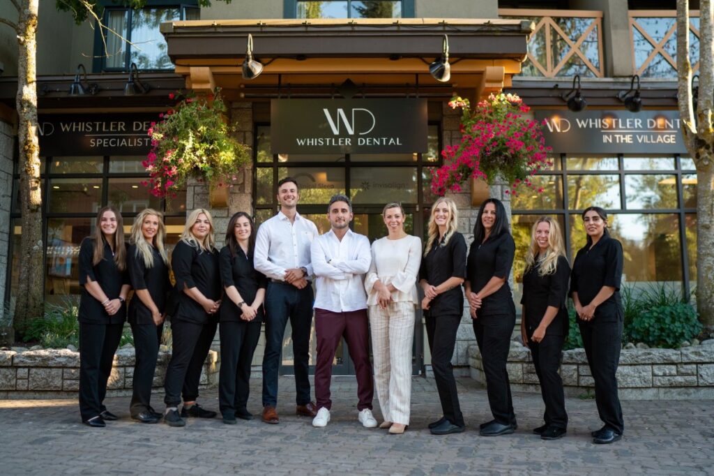 A dental team stands outside Whistler Dental, smiling and dressed in black, with two members in white attire. The welcoming atmosphere is enhanced by vibrant flowers and the modern exterior of the clinic. 123Dentist Smiley is also present, bringing a friendly touch to the scene.