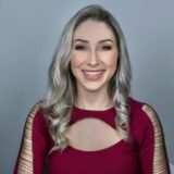 A smiling woman with long, wavy blond hair is wearing a burgundy top with cutout details. She has a friendly expression and is facing the camera against a light gray background.