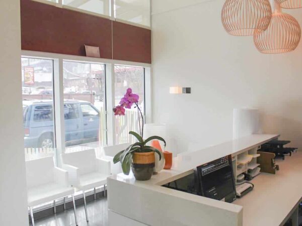 A modern reception area features a white counter, potted plants, and natural light from large windows. Stylish pendant lights hang from the ceiling, and minimalist seating is visible. The overall design is bright and inviting.