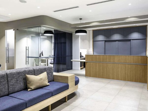 A modern office reception area featuring a comfortable gray sofa with beige cushions, an elegant wooden reception desk, and minimalist decor. Glass partitions separate workspaces, and the space is well-lit with overhead lighting.