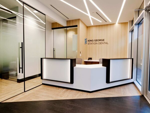 A modern dental office reception area featuring a clean design with wooden accents, illuminated signage reading "King George Station Dental," and a welcoming front desk. Vast glass walls create an open atmosphere with stylish lighting overhead.