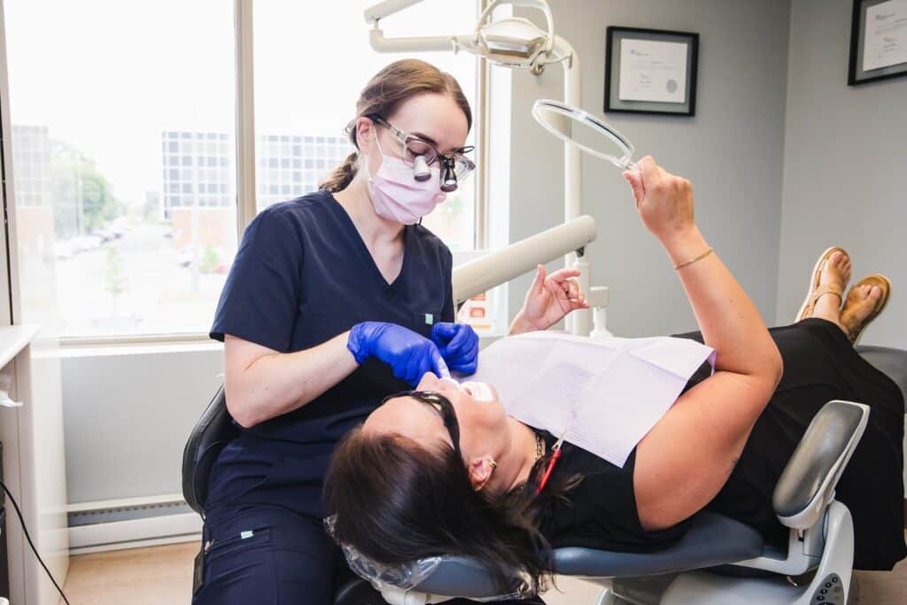 A dental hygienist works on a patient in a modern clinic, using tools to provide care. The patient lies back in the dental chair, wearing protective eyewear, while a large window reveals a view outside. The friendly 123Dentist Smiley adds a cheerful atmosphere to the setting.