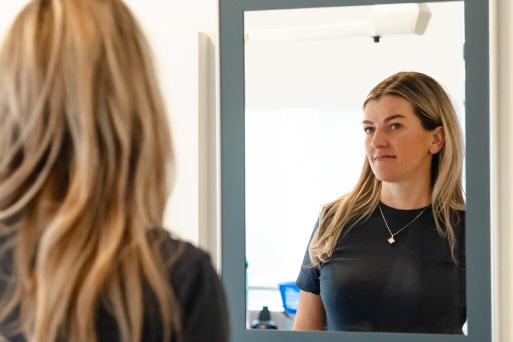 A woman with long blonde hair looks at her reflection in a mirror, displaying a thoughtful expression. The background is softly blurred, suggesting an indoor setting.