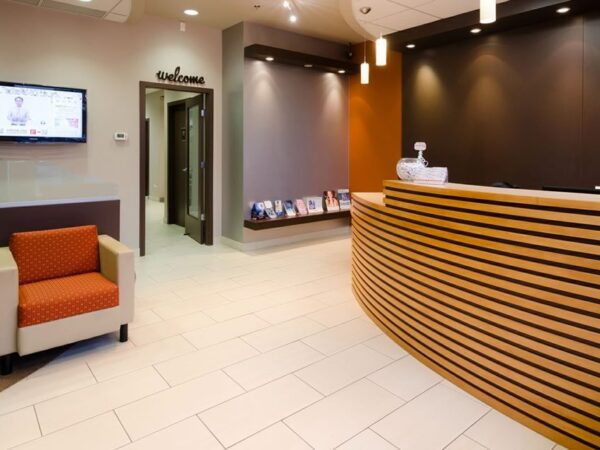 Modern lobby with a curved wooden reception desk, two orange chairs, and a wall-mounted TV. Soft lighting and neutral tones create a welcoming atmosphere.