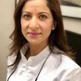 A healthcare professional wearing a white coat, with long brown hair, smiles directly at the camera. The background features a blurred image of a clinical setting, suggesting a medical environment.