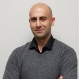 A bald man with a slight smile stands confidently against a plain background, wearing a gray sweater over a black shirt. He has his arms crossed, exuding a professional demeanor.