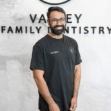 A smiling dentist wearing a black scrubs shirt stands in front of a wall with the text "Valley Family Dentistry." The background features a modern and clean aesthetic, reflecting a welcoming dental office environment.