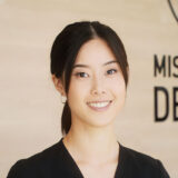 A smiling young woman wearing a black top stands in front of a light wooden background, with a sign that reads "Mission: Dentist." Her friendly expression invites a sense of warmth and approachability.