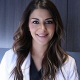 A smiling medical professional wearing a white coat over a dark top, with long, wavy hair. The background is a simple dark surface, emphasizing her friendly demeanor.