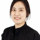 A smiling individual with black hair, wearing a black outfit, poses against a plain white background. The expression conveys friendliness and approachability.