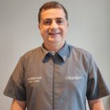 A smiling man in a gray medical coat stands against a neutral background. His coat features the name "Dr. __________ __________" and the logo of a medical institution.