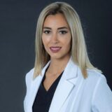 A professional woman with long, straight blonde hair is wearing a white lab coat over a black top. She is looking directly at the camera, conveying confidence and professionalism against a plain dark background.