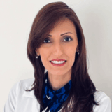 A smiling professional woman with brown hair, wearing a white coat and a blue patterned scarf. She is posed against a plain background, exuding warmth and confidence in her appearance.