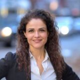 A woman with curly hair smiles confidently, standing with her hands on her hips against a blurred street backdrop with cars. She wears a black blazer over a white shirt, conveying a professional yet approachable demeanor.