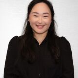 A smiling woman with long dark hair, wearing a black blouse, poses against a light gray background. She has a friendly expression, exuding warmth and approachability.