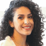 A woman with curly hair smiles warmly at the camera, wearing a white top and delicate jewelry. The background is softly blurred, focusing attention on her cheerful expression.