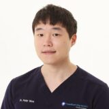 A portrait of Dr. Peter Won, wearing a dark blue dental scrubs with the logo of Coquitlam City Dentists. He has short, dark hair and is looking directly at the camera, presenting a professional demeanor.