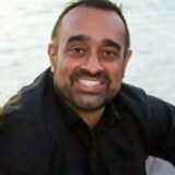 A smiling man with short black hair, wearing a dark shirt, sits by the water, with a soft, blurred background. The image captures a warm and friendly expression, conveying a relaxed atmosphere.