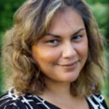 A woman with wavy, shoulder-length brown hair smiles softly. She wears a black and white patterned top and stands against a blurred green background, suggesting an outdoor setting.