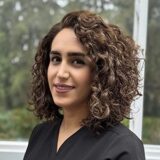 A woman with curly hair smiles at the camera. She is wearing a black top and stands in front of a window with greenery in the background, suggesting a bright and natural environment.