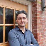 A man with short, dark hair stands confidently with crossed arms in front of a brick wall and wooden door. He is wearing a dark blue, collared shirt and has a slight smile on his face.
