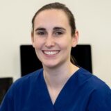 A smiling person wearing blue scrubs sits in front of a neutral background, conveying a professional and approachable demeanor.