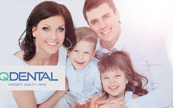 A smiling family of four, consisting of two adults and two children, is gathered closely together. The woman and man are dressed in light clothing, and the children are playfully leaning against them. The image features the logo of IQDental in the lower left corner.