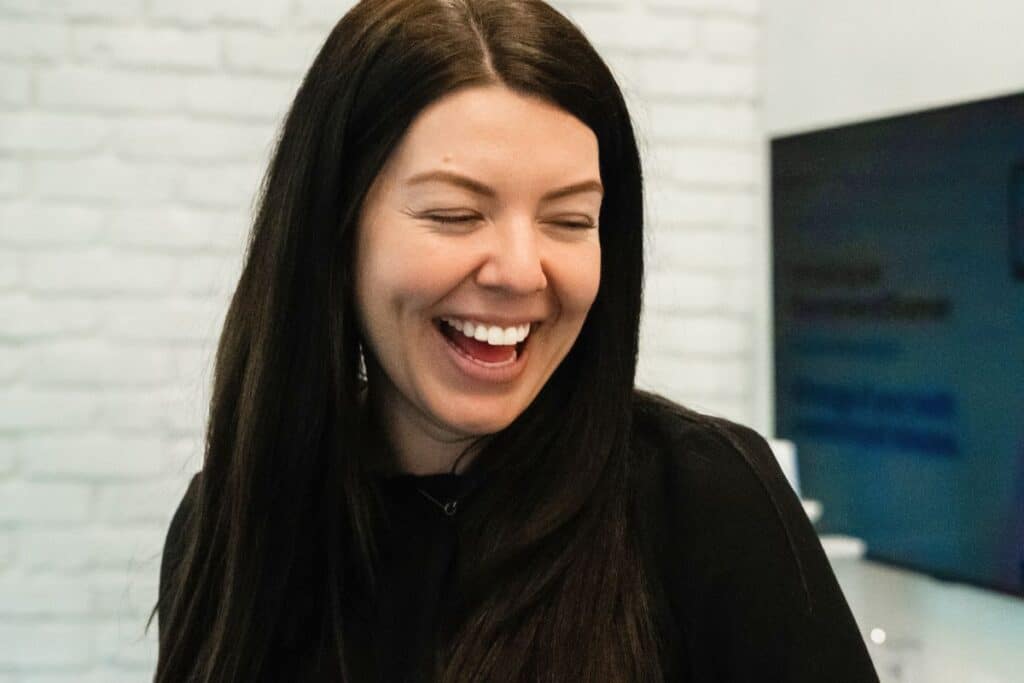 A woman with long black hair smiles joyfully, showcasing her happiness. She is dressed in a dark outfit, and her eyes are closed in delight, suggesting a warm and friendly atmosphere. In the background, a modern, minimalistic setting is visible.