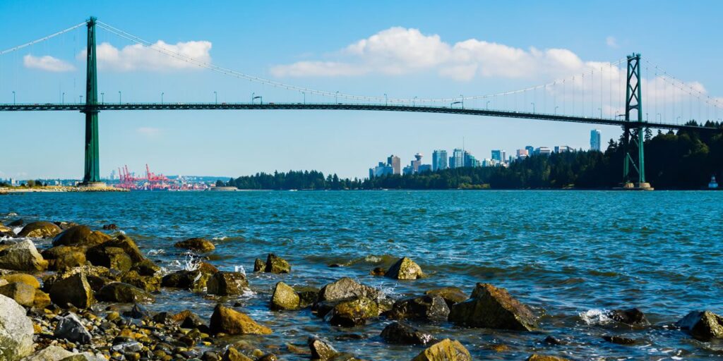 A suspension bridge spans over a blue body of water, with rocky shores in the foreground and a city skyline in the background under a clear sky.