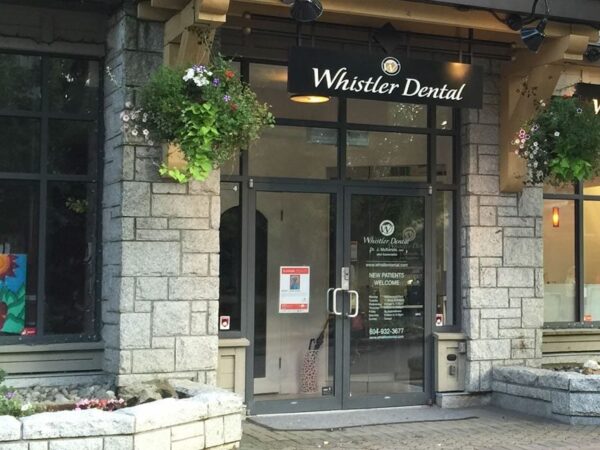 Front entrance of Whistler Dental, featuring a glass door, large windows, stone walls, and potted plants.