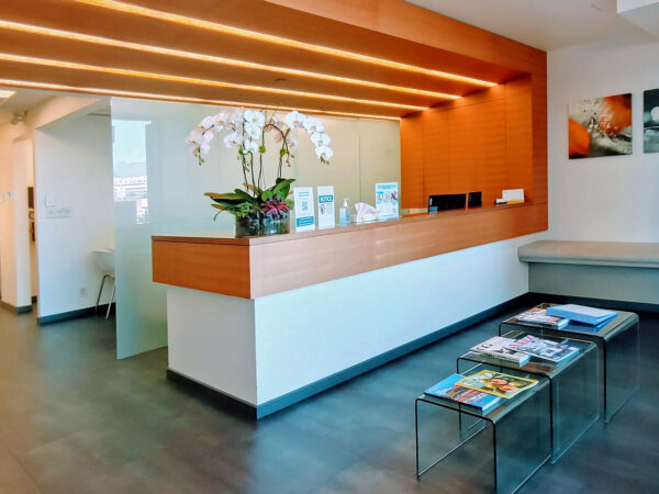 A modern dental office reception area featuring a sleek white front desk with a floral arrangement, warm wood accents, and a seating area with magazines on a glass table. The space is bright and welcoming, designed for patient comfort.