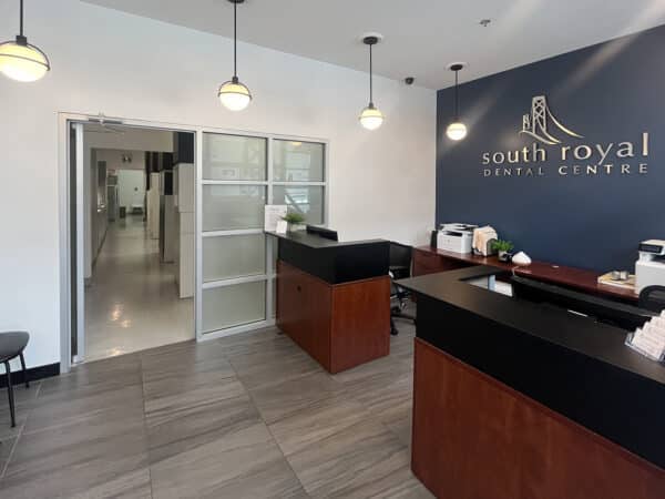 A modern dental office reception area with a brown counter, a welcoming atmosphere, and branding for South Royal Dental Centre on the wall. The space features stylish lighting and waiting chairs, creating an inviting environment for patients. The friendly 123Dentist Smiley adds a touch of warmth.