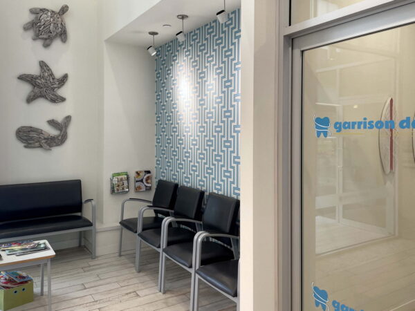 A modern waiting area featuring a bench with metal chairs, a blue patterned accent wall, and decorative bird sculptures. The space is well-lit and includes a glass door labeled with "garrison dental." Magazines are neatly arranged on a small table.