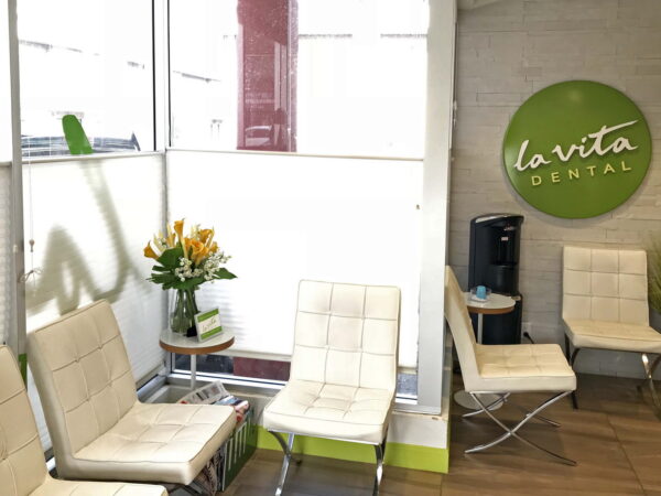 A modern dental clinic waiting area featuring white chairs, a small table with a vase of flowers, and a wall sign that reads "la vita dental." Natural light filters through large windows, creating a bright and inviting atmosphere.