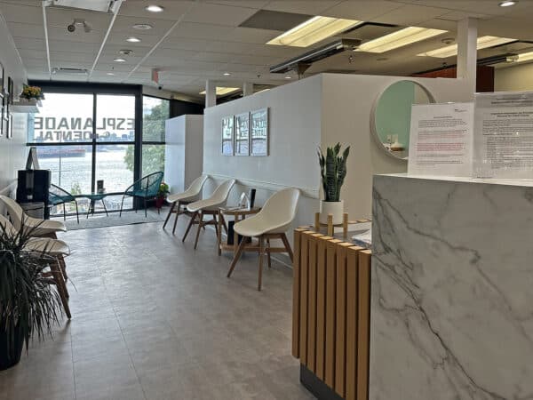 A modern dental office reception area featuring a spacious waiting room with wooden chairs, potted plants, and large windows. The front desk, made of marble, is neatly organized with informational materials visible. 123Dentist Smiley adds a friendly touch to the inviting environment.