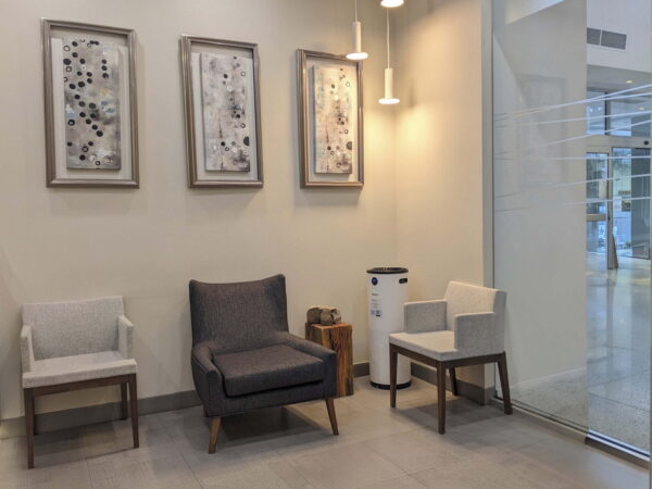 A modern waiting area featuring three chairs, including a dark gray armchair, complemented by two light-colored chairs. The walls display three abstract artworks, and a trash can is positioned next to a wooden side table. Natural light floods the space through large glass windows.